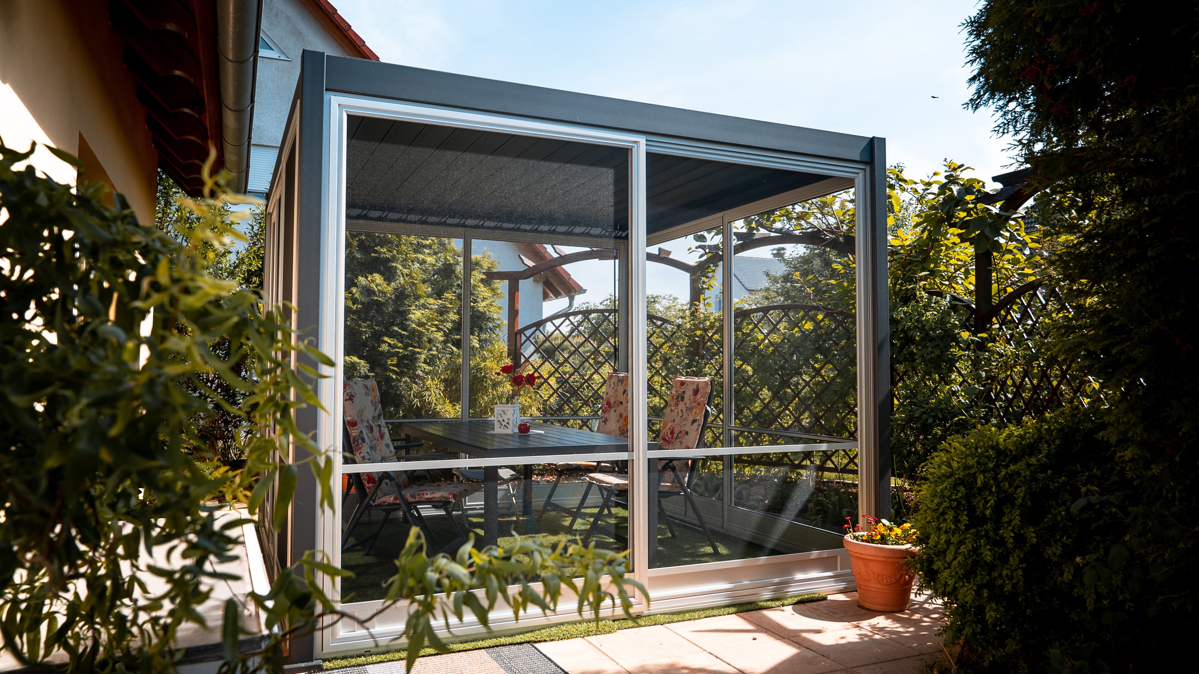 Ein Pavillon mit Insektenschutz-Gitter welche sich verschieben lassen, welcher auf der Terrasse steht. Darin befinden sich 4 Stühle und ein Tisch. Es ist ein sonniger Tag, die Bäume sind Grün.